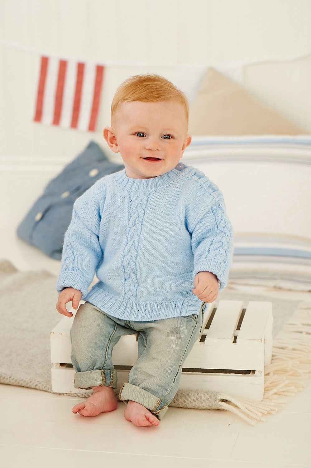 A baby is wearing a light blue knitted sweater with cable down the front centre.