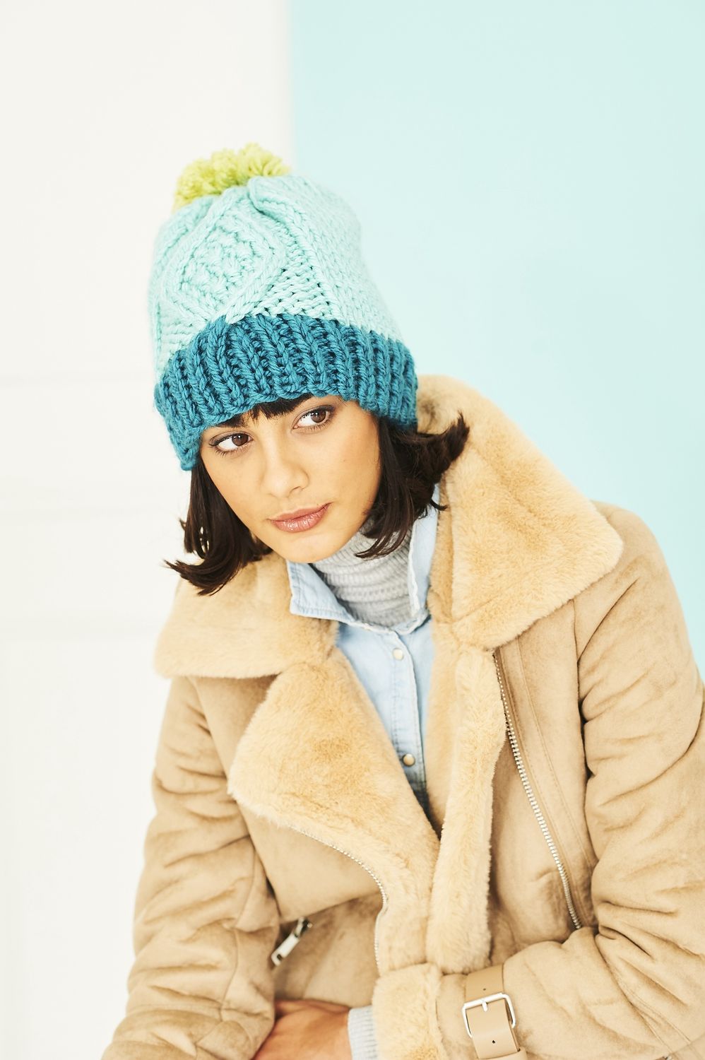 A woman is wearing a light blue cable knit hat, with a dark blue brim and a green pompom.