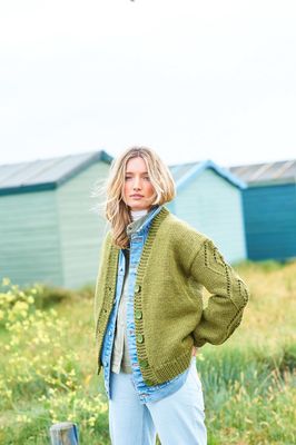 A woman is standing in a field in front of a row of blue beach huts, wearing a green cardigan with lace detail on the sleeves.