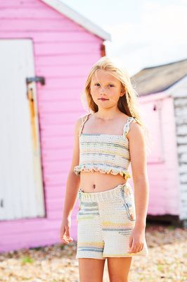 A girl is wearing a crocheted top and shorts in shades of cream, grey, green, peach and yellow.