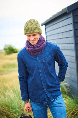 A man is standing in a field in front of a dark blue building, wearing a green hat and a purple snood.