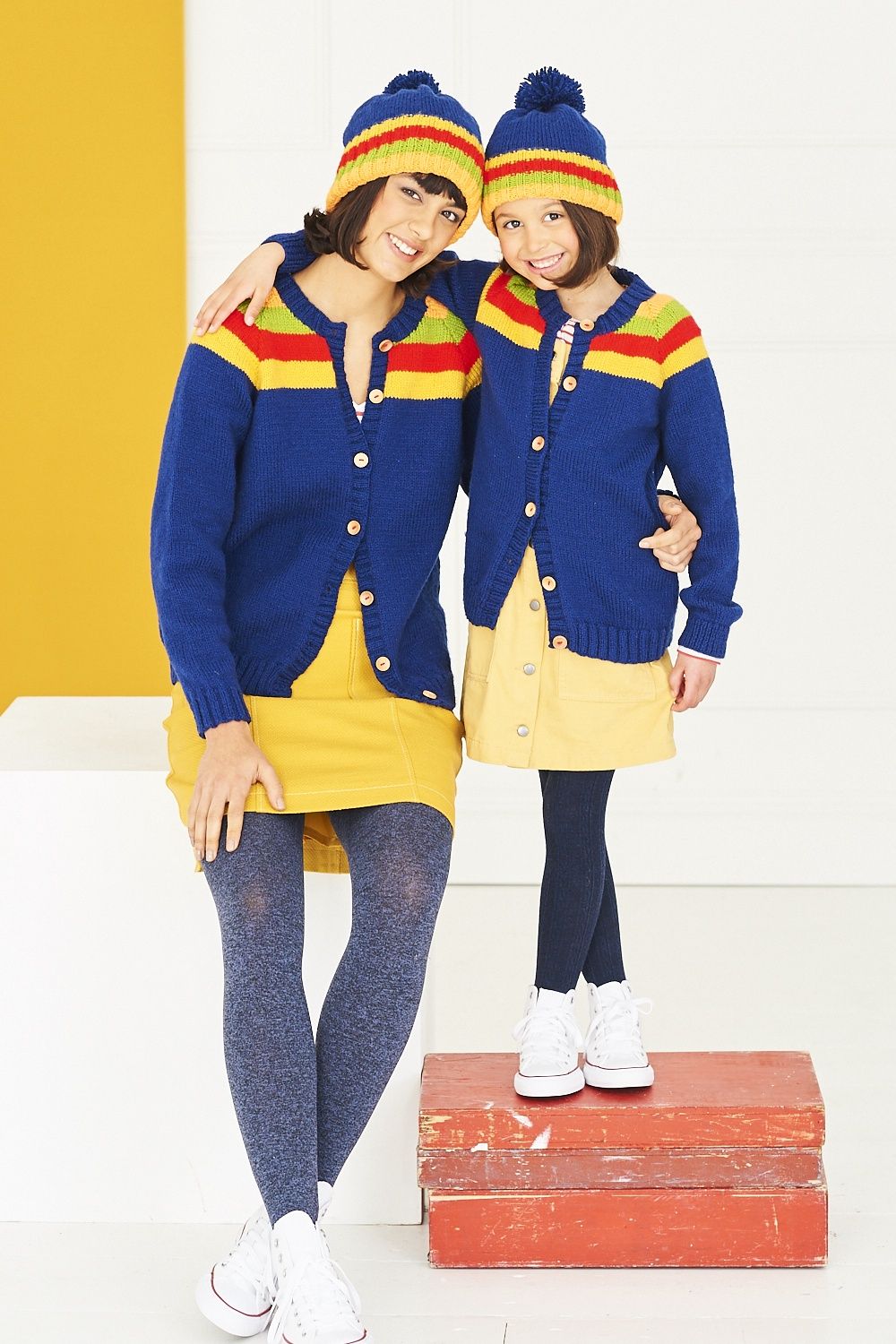 A woman and a girl are wearing matching navy blue knitted cardigan with yellow, green and red stripes.