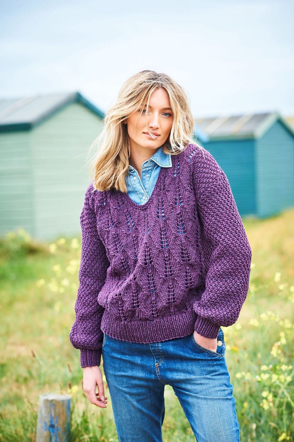 A woman is standing in a field in front of a row of blue beach huts, wearing a dark purple sweater with lace detail on the front and textured sleeves.