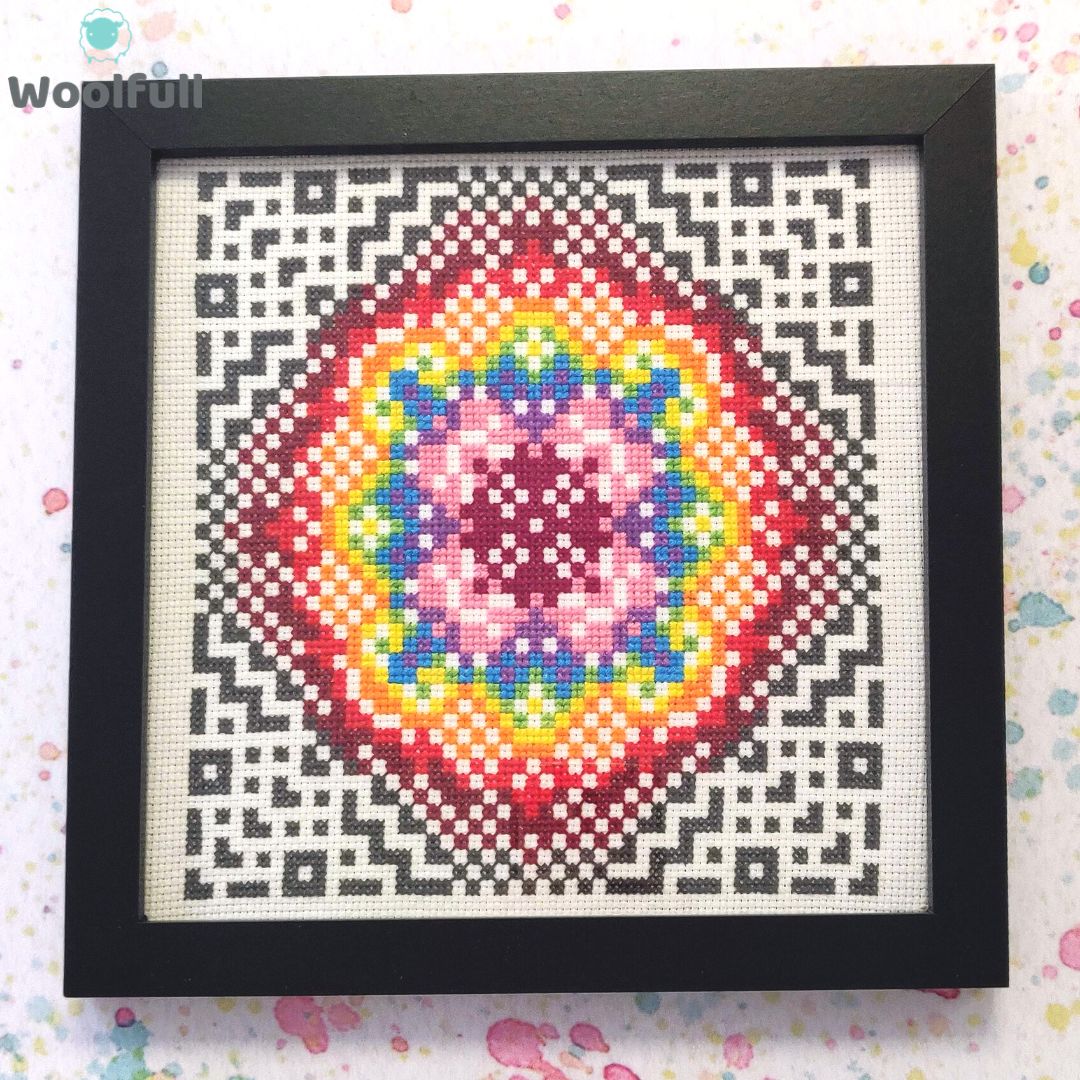 A framed cross stitch with a design of a rainbow coloured geometric mosaic surrounded by grey geometirc shapes and lines.