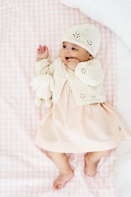 A baby is wearing a cream knitted cardigan and matching hat.