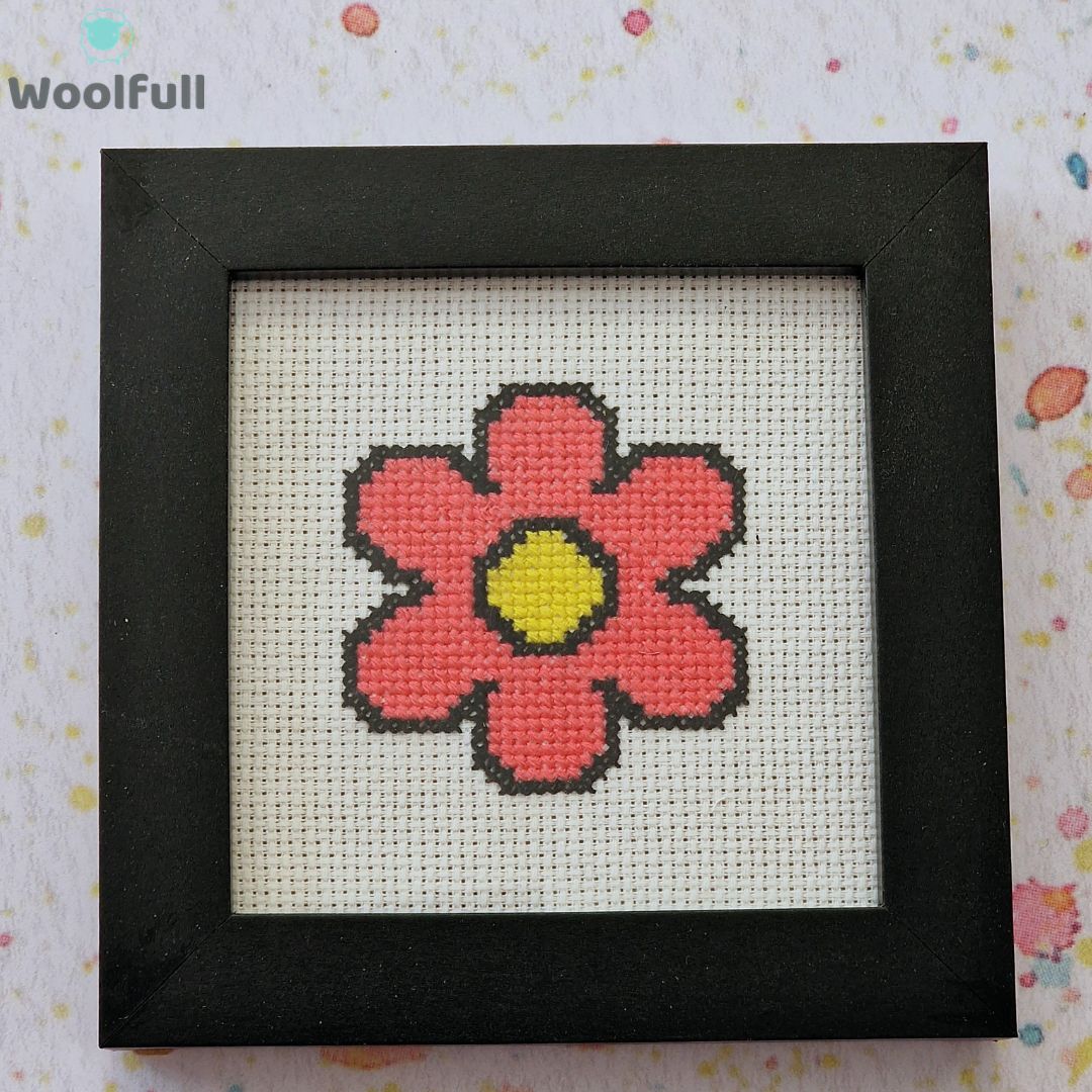 A framed cross stitch with a design of a pink cartoon style flower.