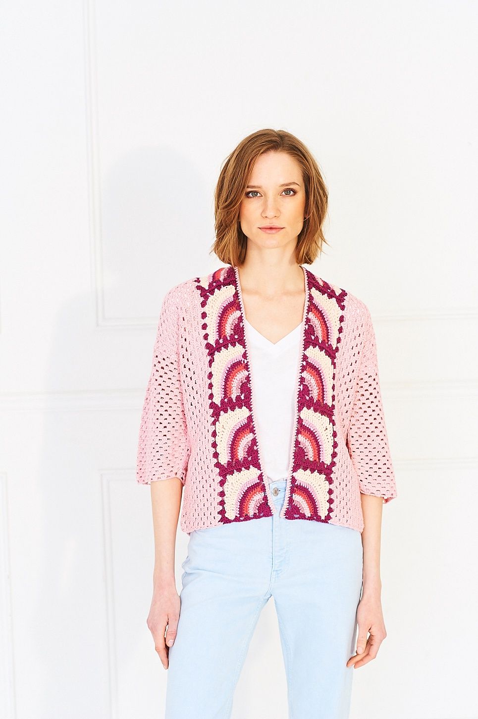 A woman is wearing a pink crocheted cardigan with rainbow granny squares on the front and back.