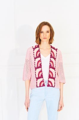 A woman is wearing a pink crocheted cardigan with rainbow granny squares on the front and back.