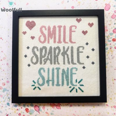 A framed cross stitch with a design that says &#39;smile, sparkle, shine&#39; in pink, silver and blue.