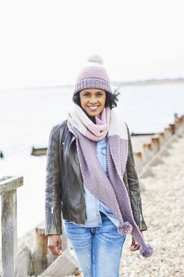 A woman is wearing a knitted scarf and matching hat in cream, pink and purple.