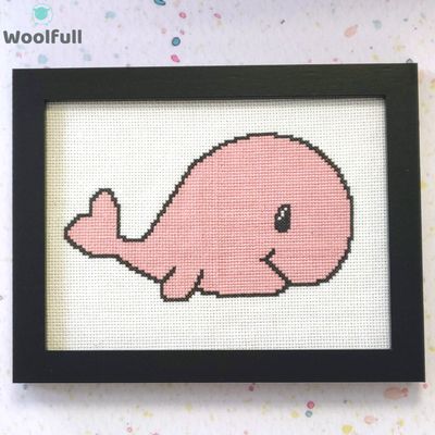 A framed cross stitch with a design of a pink cartoon style whale.