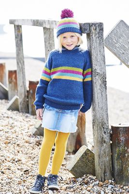 A girl is wearing a dark blue knitted sweater with green, magenta, aqua and yellow stripes, and a matching hat with a magenta pompom.