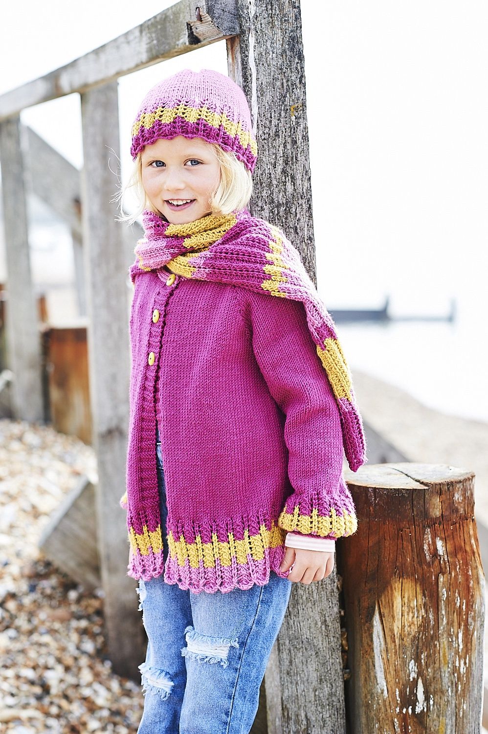 A girl is wearing a pink and yellow knitted cardigan, with matching hat and scarf.