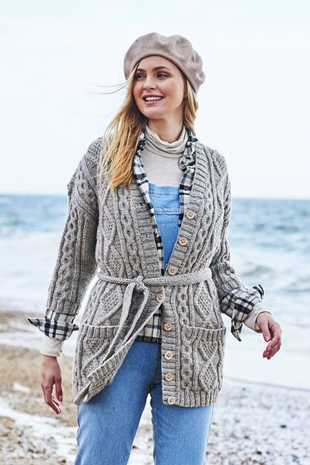 A woman on the beach is wearing a long grey cabled cardigan with two pockets on the front.