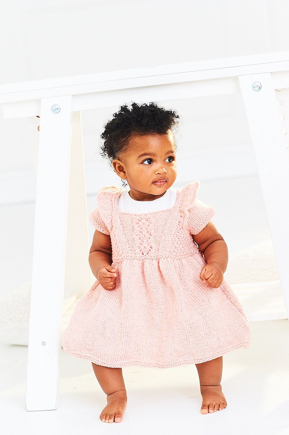 A baby is wearing an apricot coloured knitted dress.