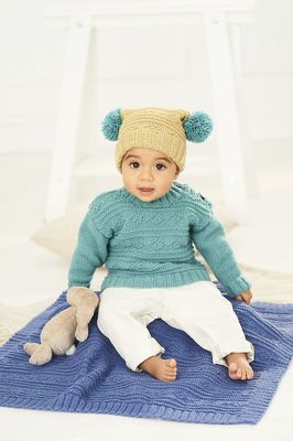 A baby is sitting on a blue knitted blanket, while wearing a light teal knitted sweater and a light green knitted hat with teal pompoms.