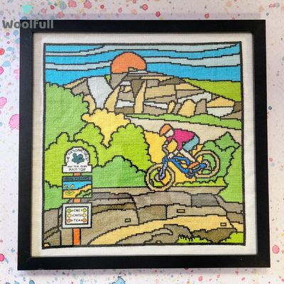 A framed cross stitch with a cartoon style design of a cyclist on Mam Tor in the Peak District.
