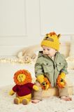 A baby is wearing a yellow knitted lion hat and matching mittens, while sitting next to a knitted toy lion in a red sweater.