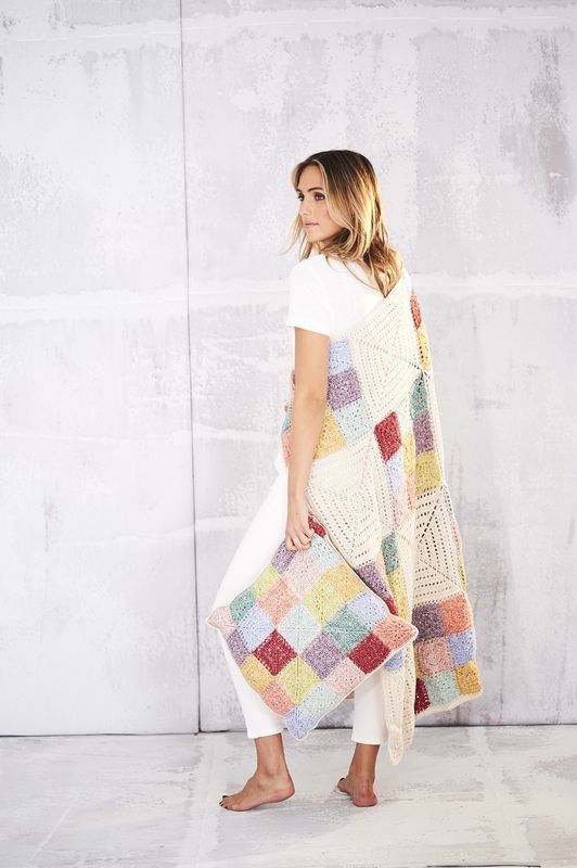 A woman is wearing a crocheted blanket made up of cream, purple, pink, yellow, blue, green and coral squares and holding a matching cushion.
