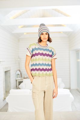 A woman is wearing a knitted top with a wavy stitch pattern in cream, purple, blue and green with a hat in matching colours.