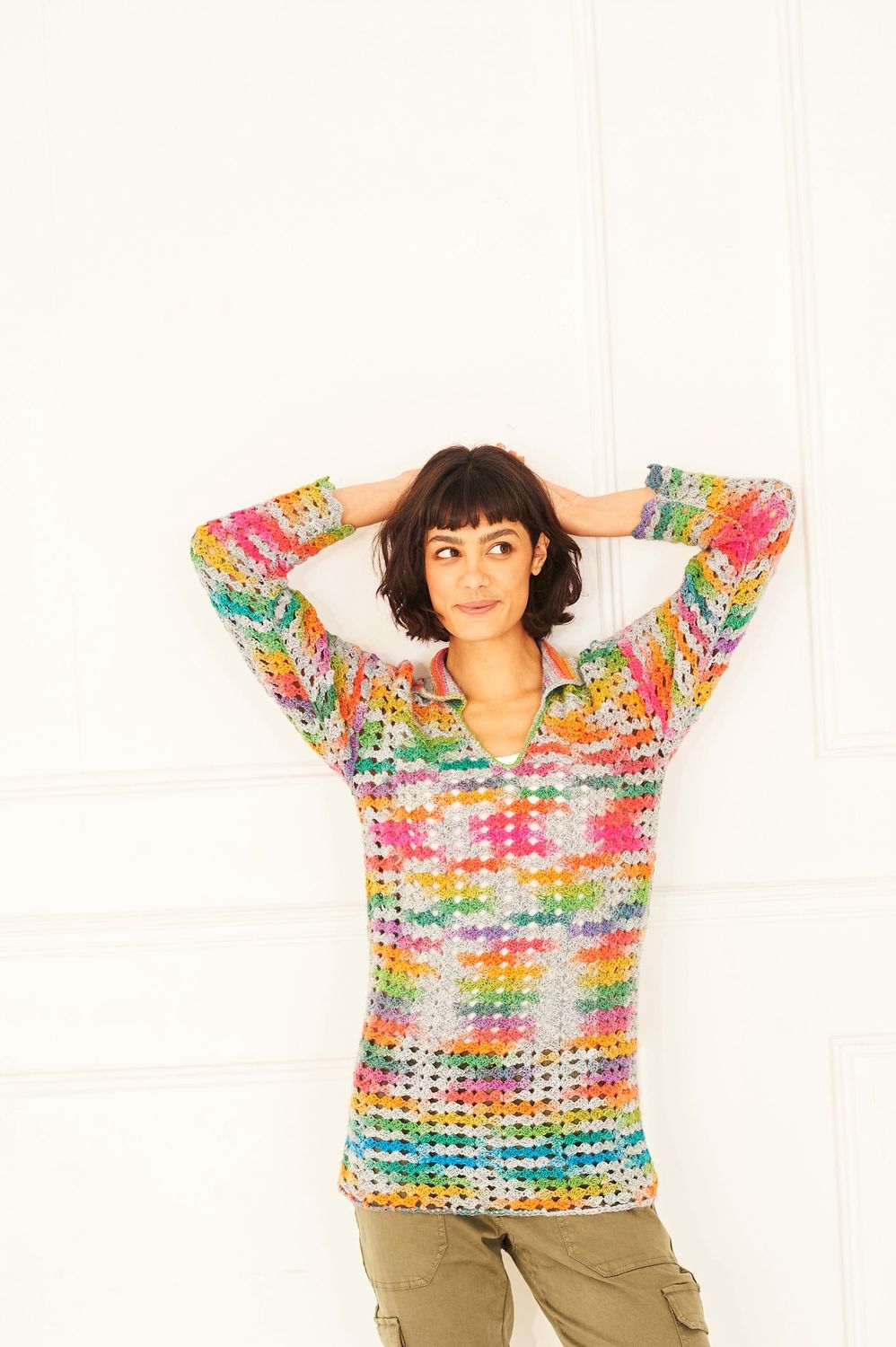 A woman is wearing a crocheted tunic in shades of grey, orange, yellow, green, blue and purple and pink.