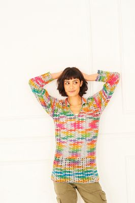A woman is wearing a crocheted tunic in shades of grey, orange, yellow, green, blue and purple and pink.