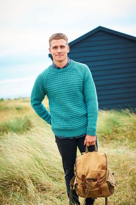 A man is standing in a field in front of a dark blue building, wearing a teal coloured textured sweater.