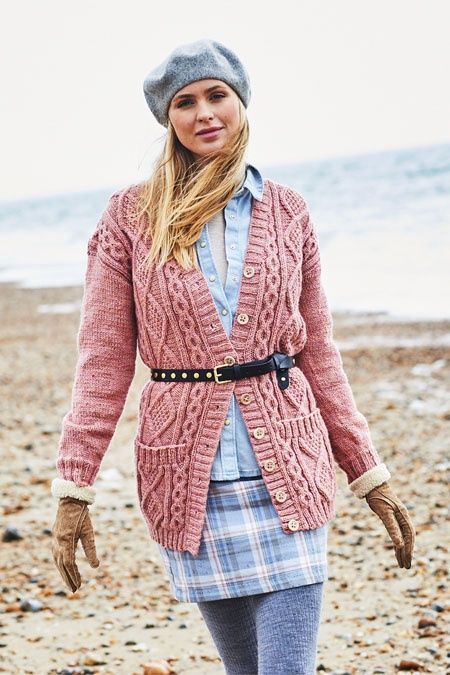 A woman on the beach is wearing a long pink cabled cardigan with two pockets on the front.