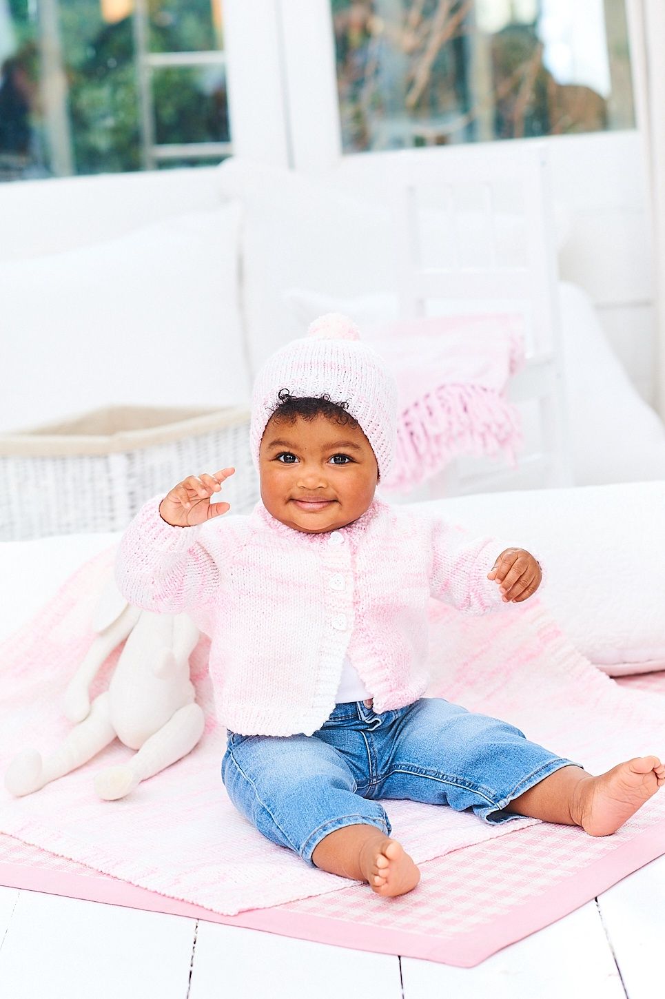 A baby is wearing a pink knitted cardigan with a matching hat and blanket.