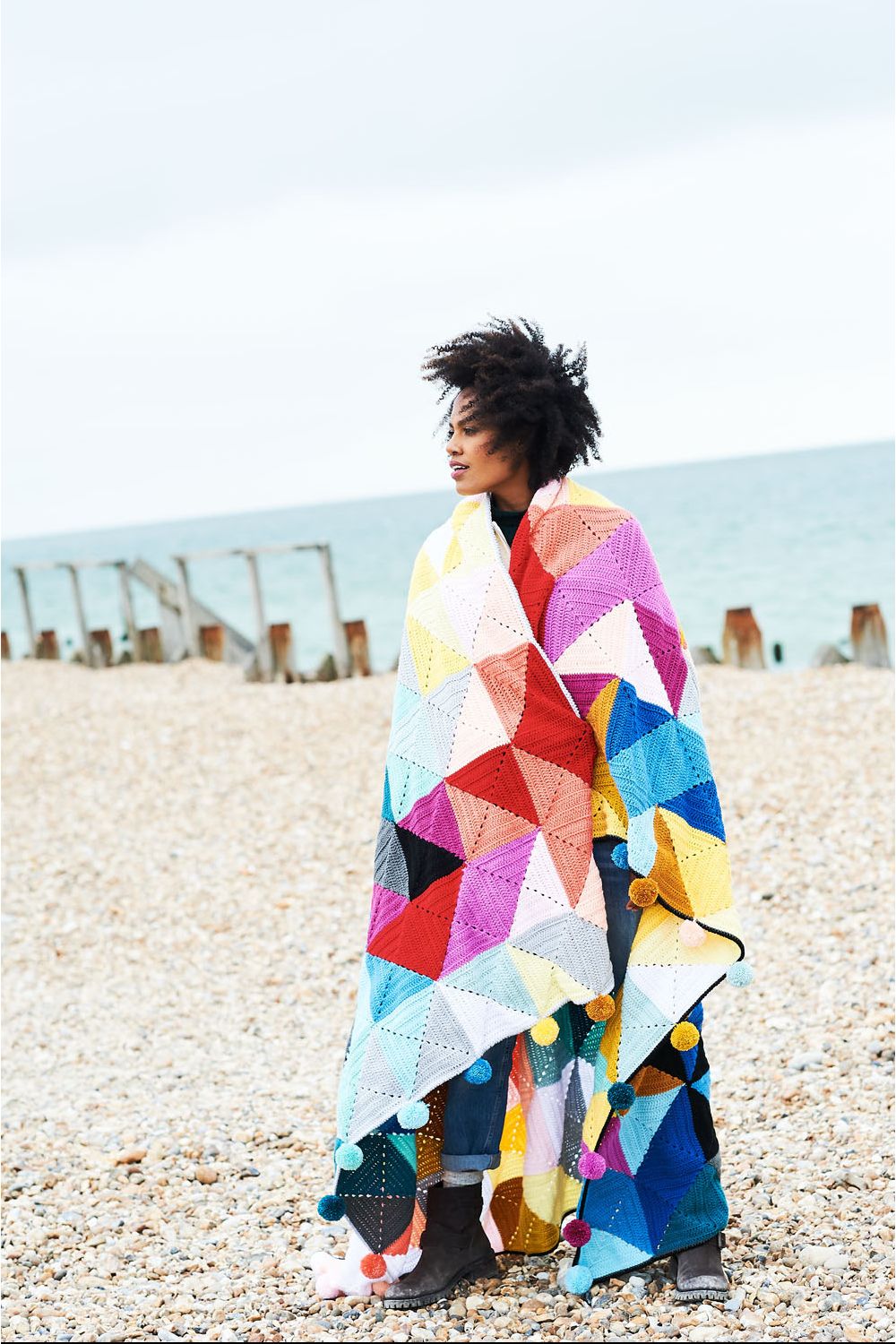 A woman has a crocheted blanket in shades of white, pink, red, blue and yellow draped around her shoulders.