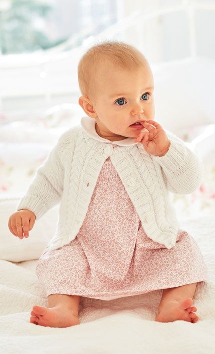 A baby is wearing a white cardigan with cable detail on the front.