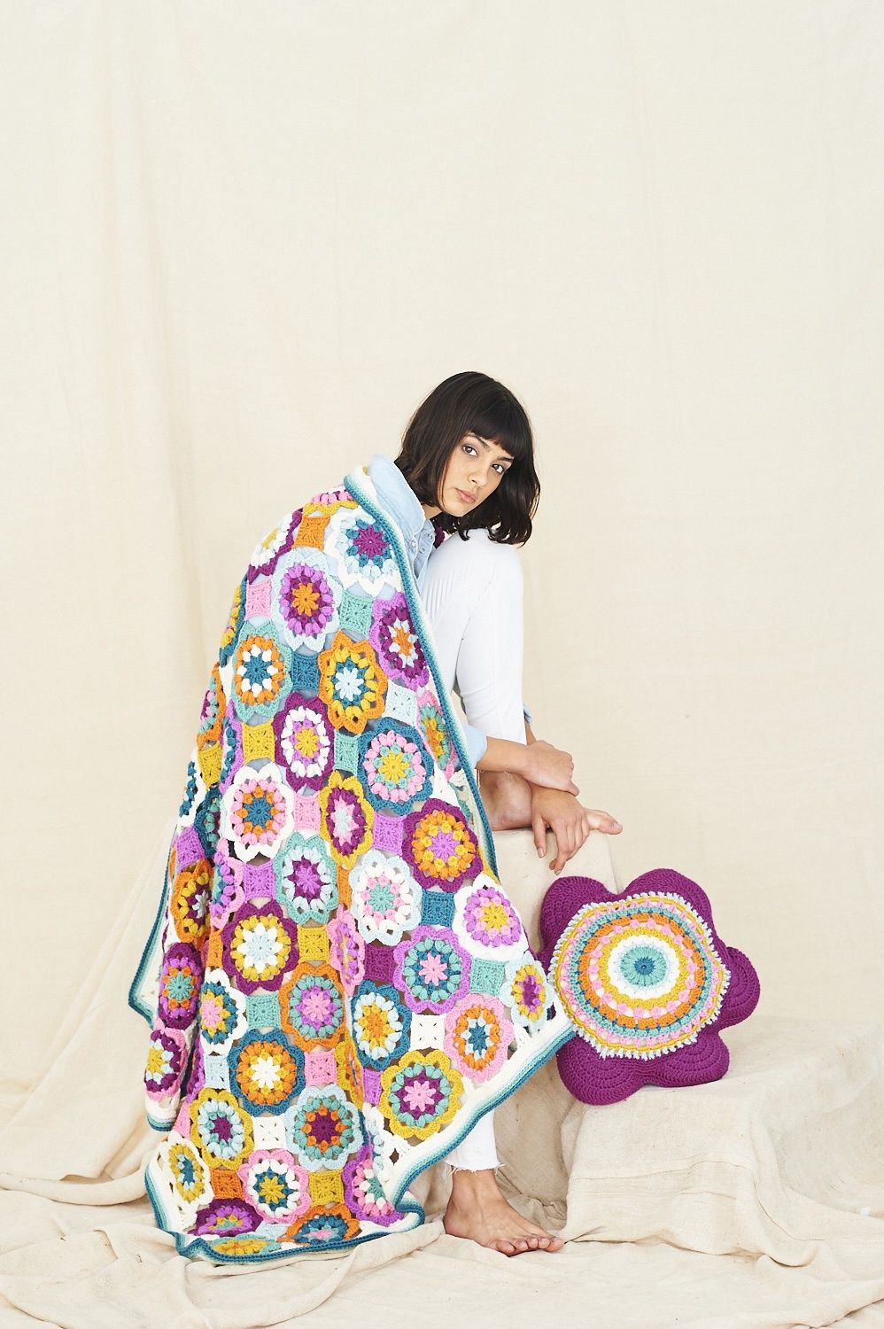 A woman is wearing a crocheted blanket made up of pink, purple, blue, orange and yellow flower shapes.