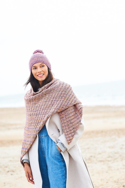 A woman is wearing a knitted hat and shawl in shades of pink, purple, yellow and brown.