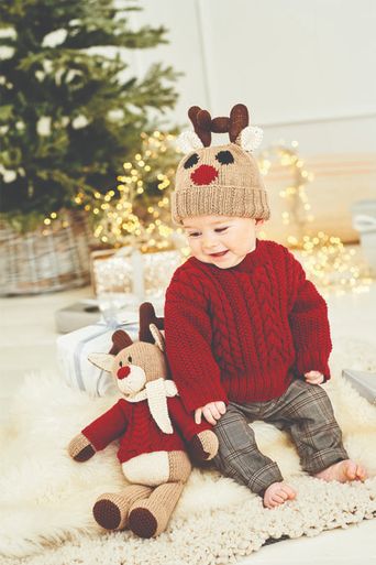 A baby is wearing a red cable knit sweater and a brown reindeer hat, while sitting next to a knitted reindeer toy.