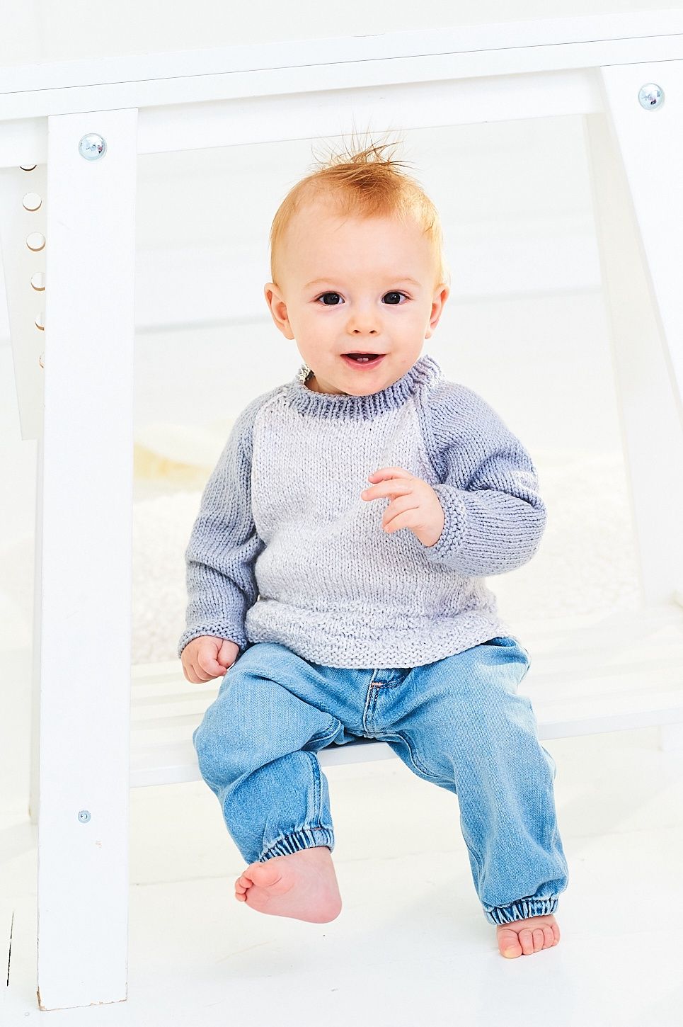 A baby is wearing a knitted sweater in light grey and mid grey.