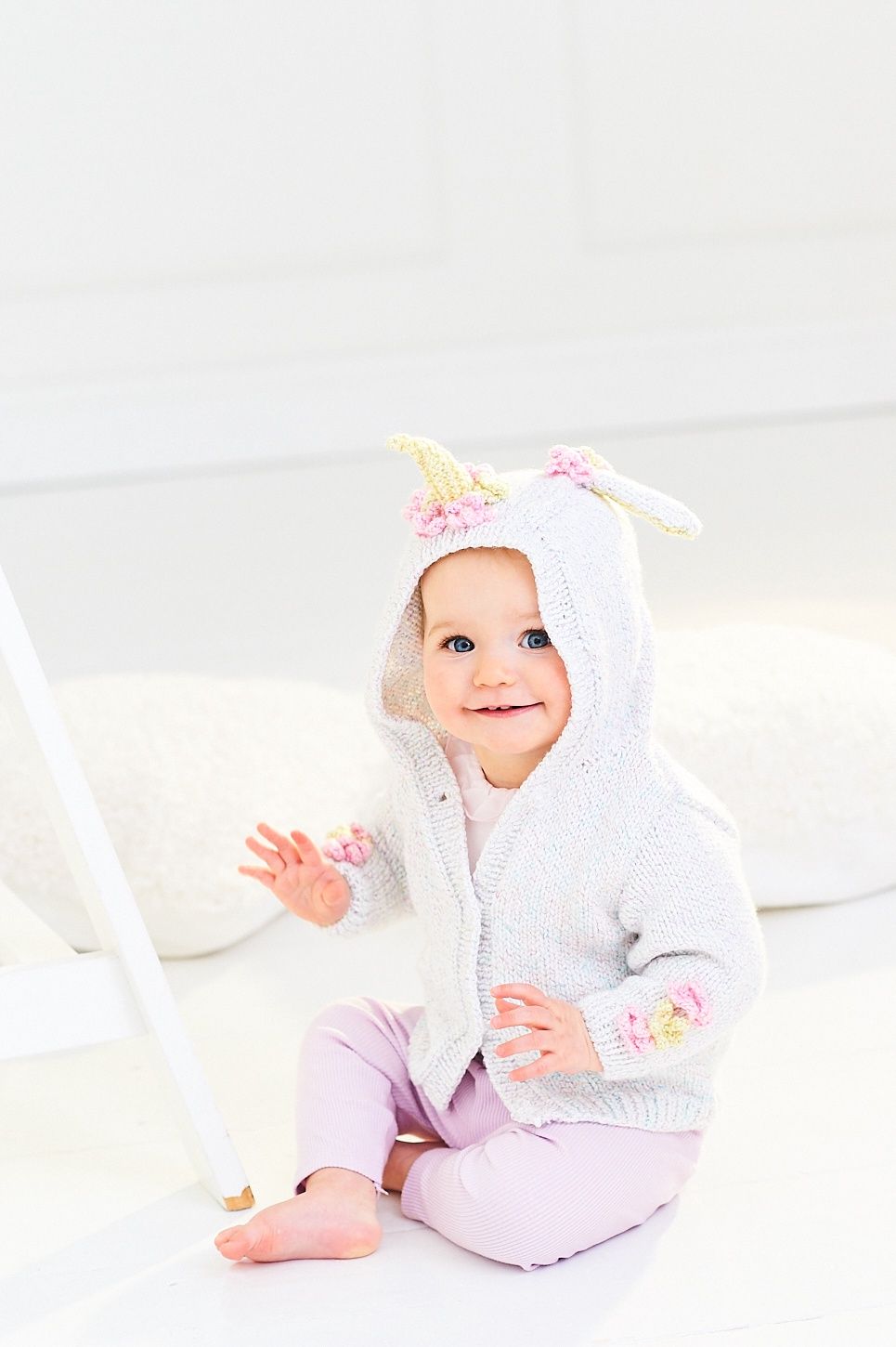 A baby is wearing a knitted hooded unicorn cardigan in white, pink and yellow.