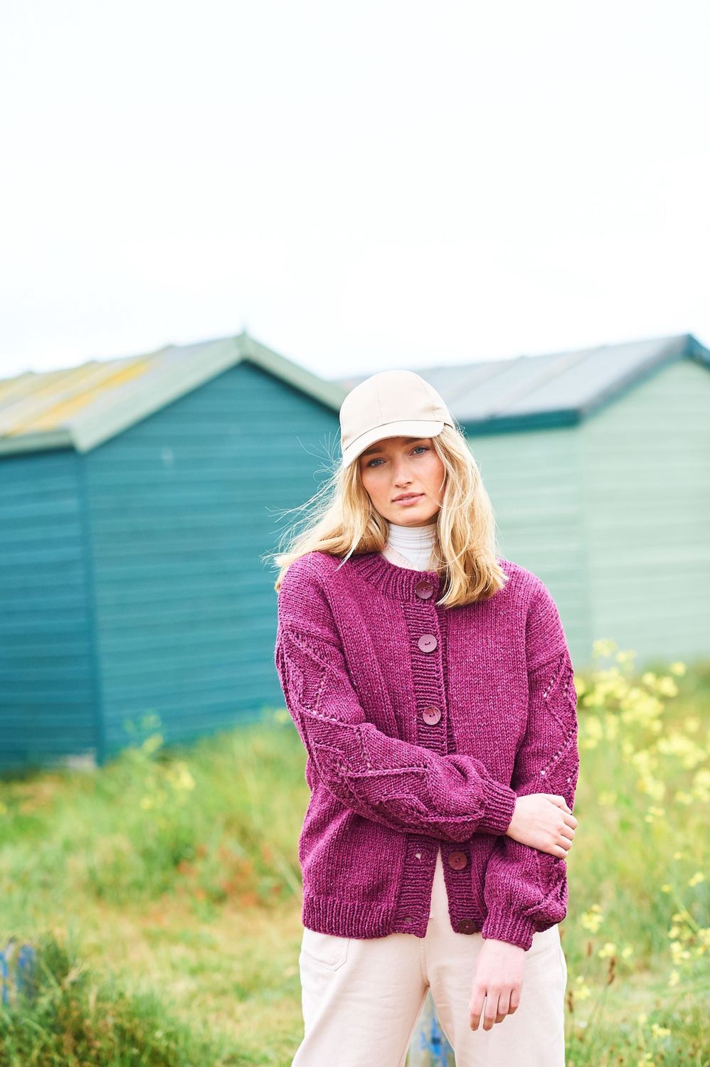 A woman is standing in a field in front of a row of blue beach huts, wearing a dark pink cardigan with lace detail on the sleeves.