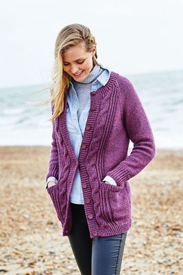 A woman on the beach is wearing a long purple cardigan with cable details down the front.