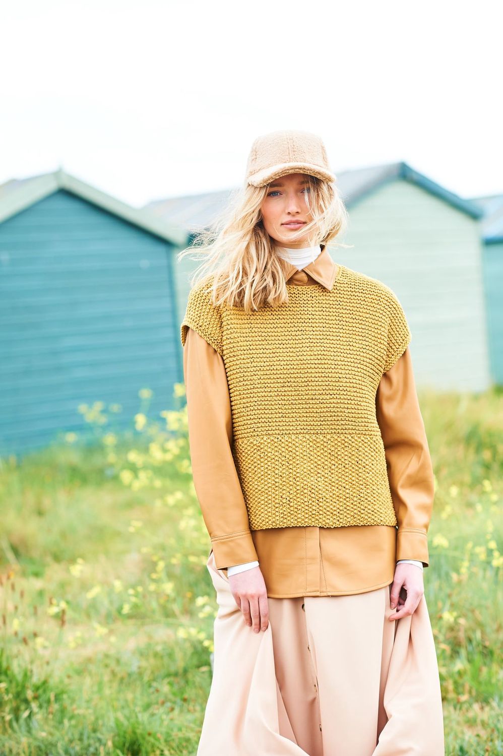A woman is standing in a field in front of a row of blue beach huts, wearing a mustard coloured tank top over an orange shirt.