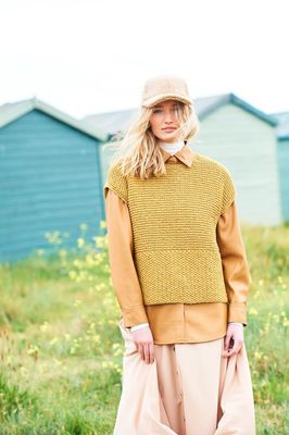 A woman is standing in a field in front of a row of blue beach huts, wearing a mustard coloured tank top over an orange shirt.