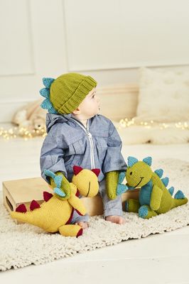A baby is wearing a green knitted hat with blue spikes and matching mittens, while sitting next to 2 knitted dinosaur toys.