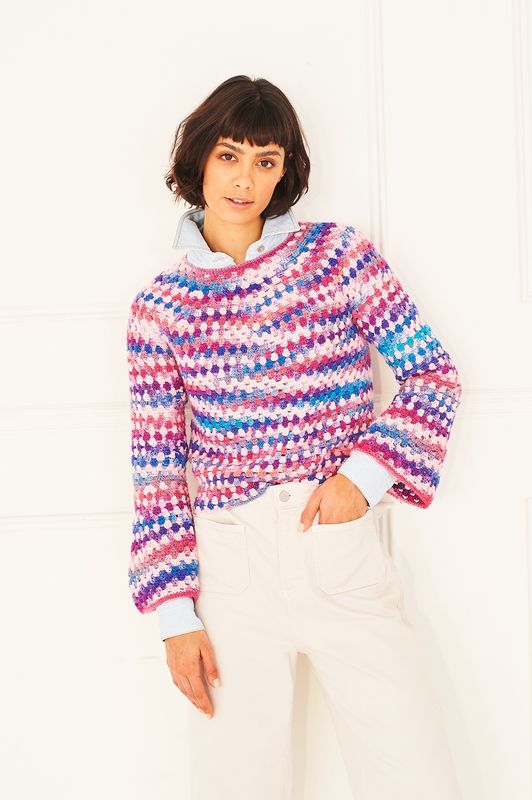 A woman is wearing a crocheted sweater in shades of pink, blue and purple.