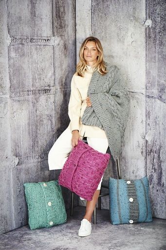 A woman is wearing a grey knitted blanket, holding a pink knitted cushion and has two other knitted cushions by her feet.