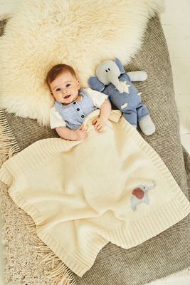 A baby is under a cream knitted blanket with an elephant motif, next to a blue and grey knitted elephant toy.