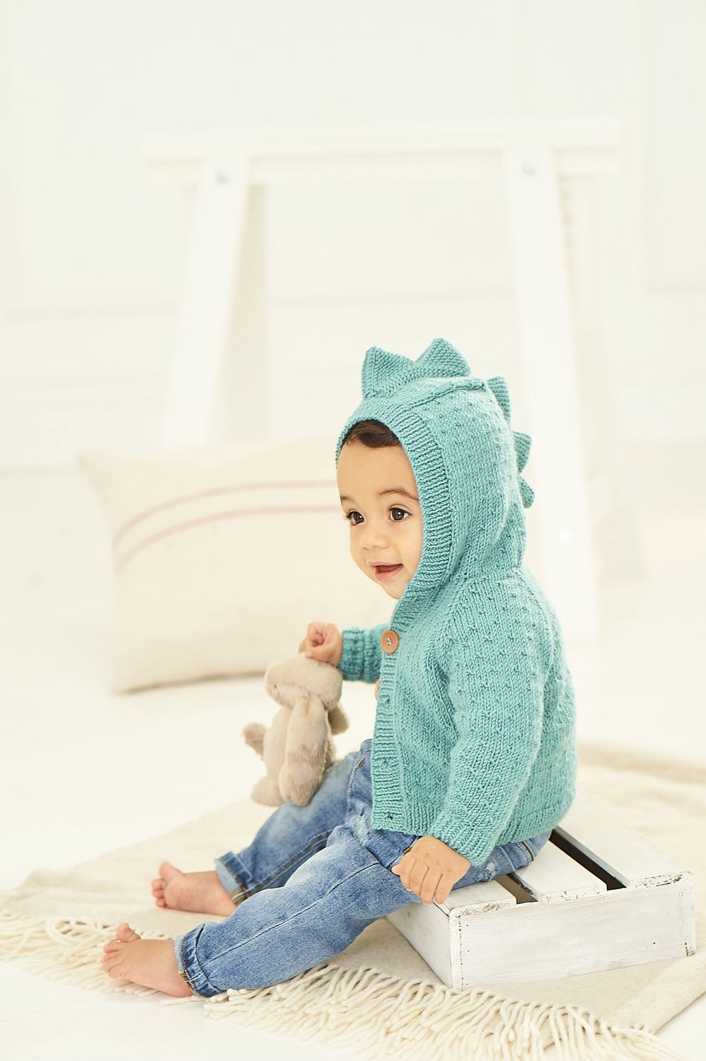 A baby is wearing a teal knitted hooded cardigan with dinosaur spikes down the hood and back.