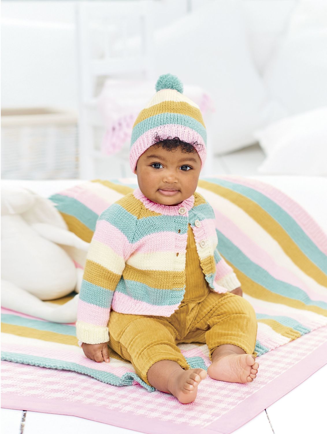 A baby is wearing a striped cream, pink, blue and yellow knitted cardigan with a matching hat and blanket.