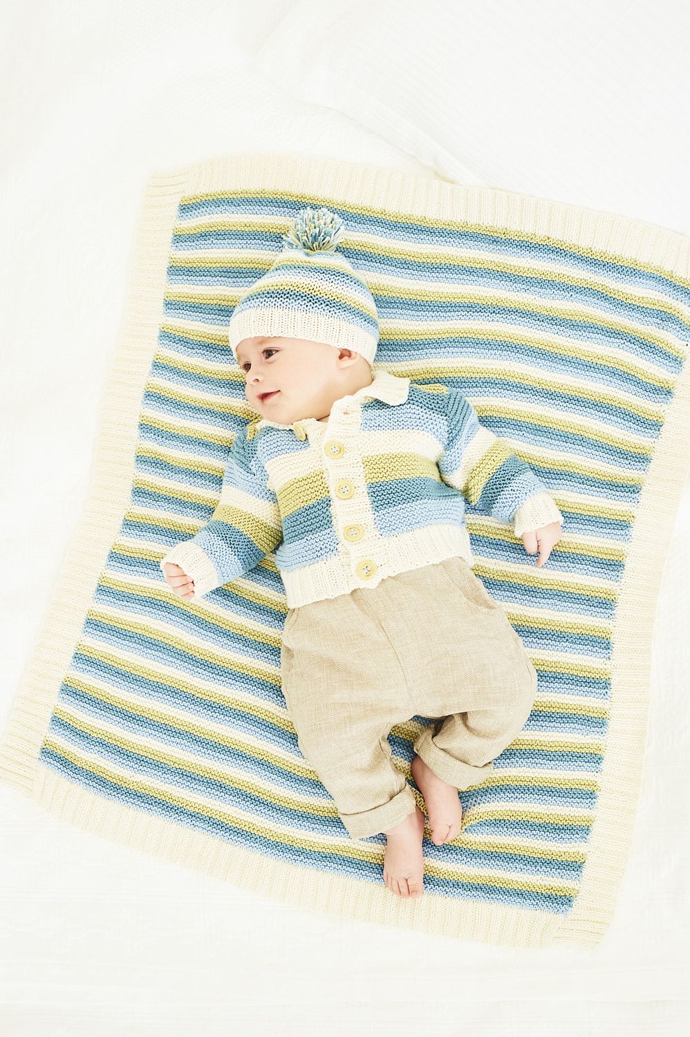 A baby is wearing a knitted cardigan and matching hat in blue, green and cream stripes, while laying on a matching blanket.
