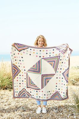 A woman is holding a crocheted blanket made up of squares in cream and various bright colours.