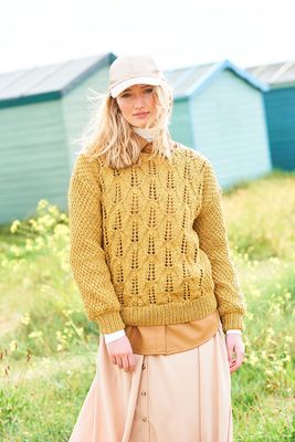 A woman is standing in a field in front of a row of blue beach huts, wearing a mustard yellow sweater with lace detail on the front and textured sleeves.