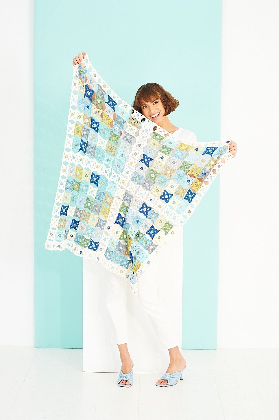 A woman is holding a crocheted blanket made up of white, blue, green, grey and yellow squares.